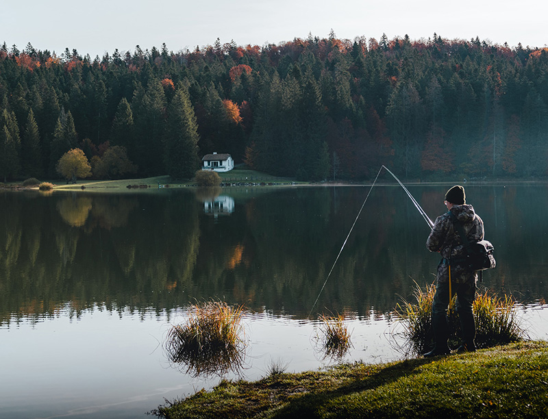 A person in camouflage with a backpack and fishing pole, fishing on the calm waters of the lake. A pine forest and a small cozy hut stand on the opposite bank.