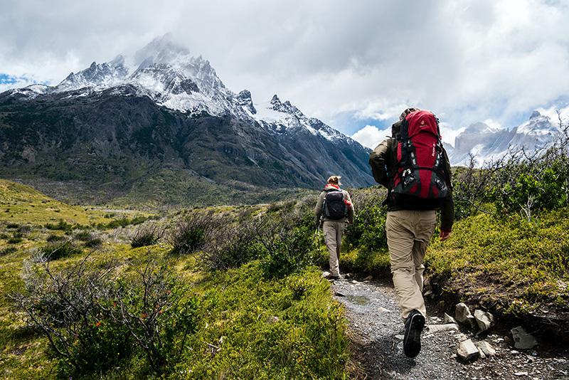 Two people with backpacks hiking along a narrow mountain path, en route to the snow-covered peaks.