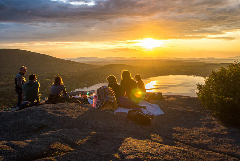 A group of people sitting on a rock, looking down at a lake during the sunset.